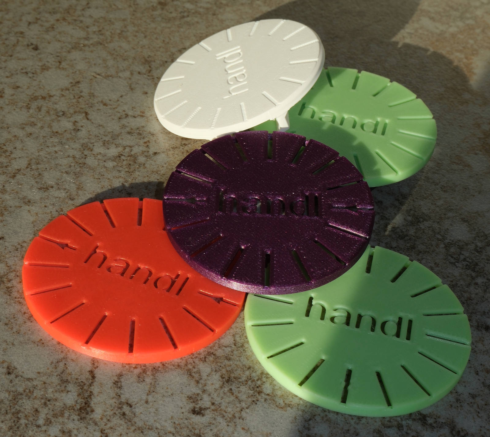 A photograph of five real printed parts laid on a speckled worktop in sunlight: four coloured PALM clips — red, dark purple and two greens — and, at the top, one white adapter. The clips are cut through at the rim so daylight shows in the slots; the white adapter has an unbroken edge. Colours photographed in hard sun, not colour swatches.